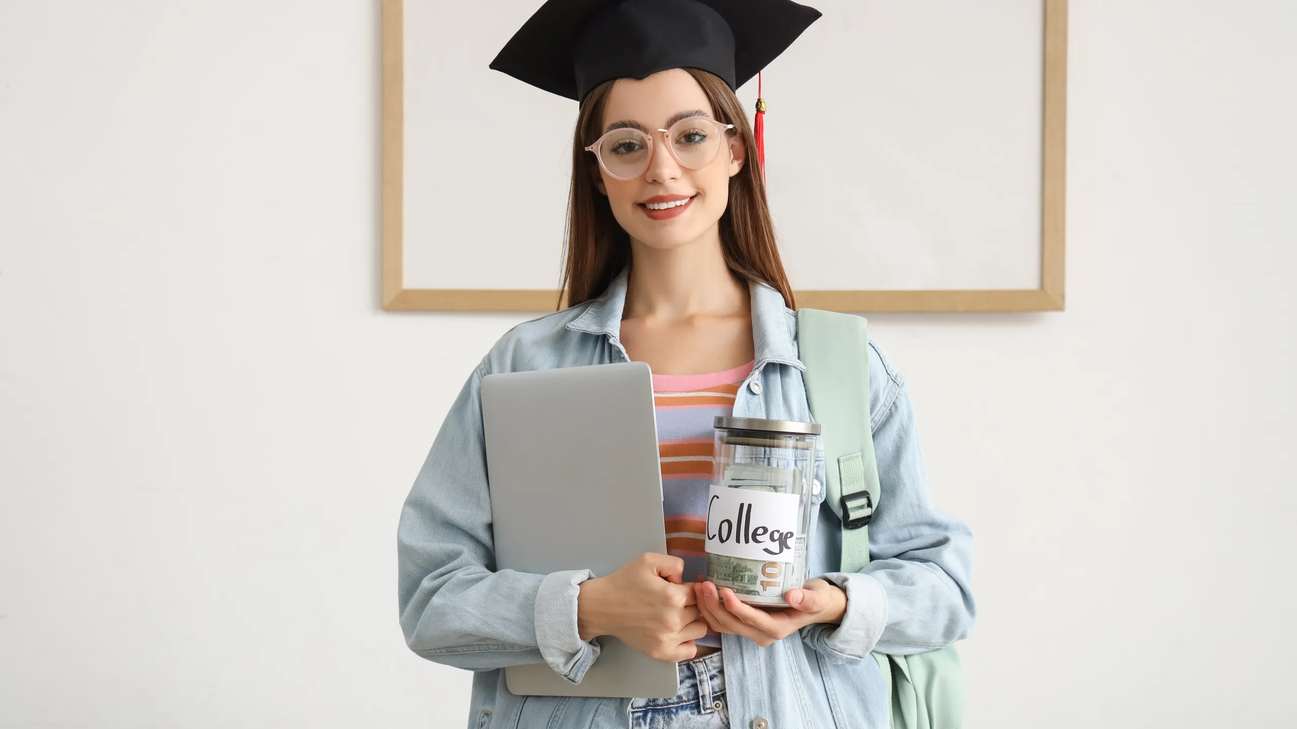 Female student in graduation hat with laptop and jar of money for college at home. Education savings concept