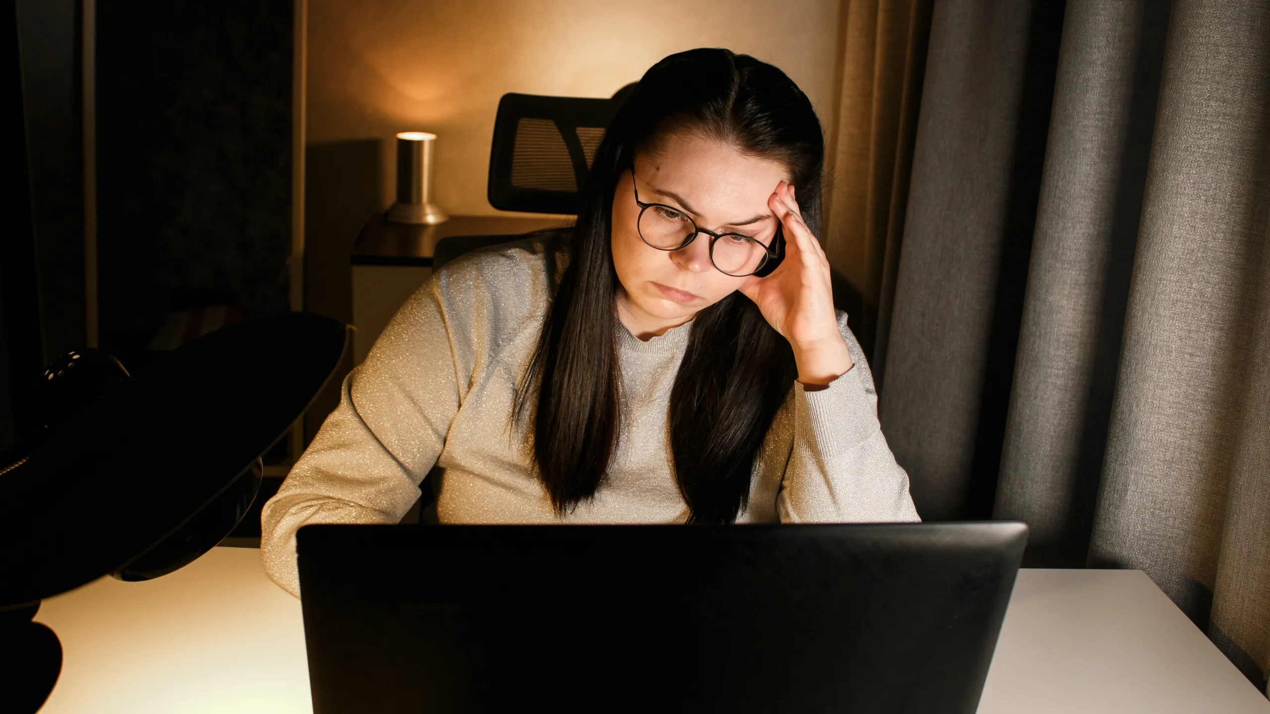 Young woman with eyeglasses working on laptop at home late at night, using modern technologies and getting online education. Remote learning concept. Online university. Focus on process.