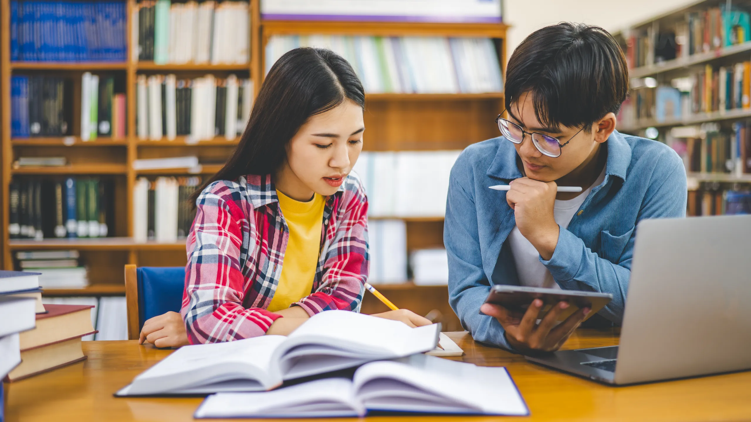 Students are consulting each other working on laptops and searching for books to study, make reports, find useful information in university room. Concept of reading, learning and intelligence.