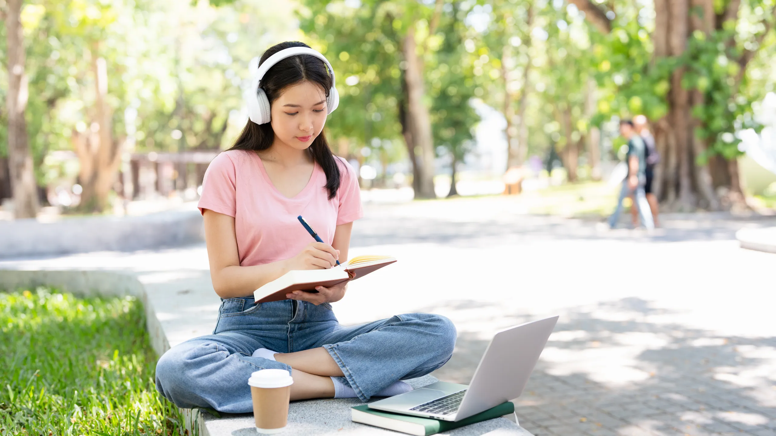 Smiling young asian woman in wireless headphones sitting outdoor working on laptop computer and writing or taking notes in paper notebook, holding pen in hand.