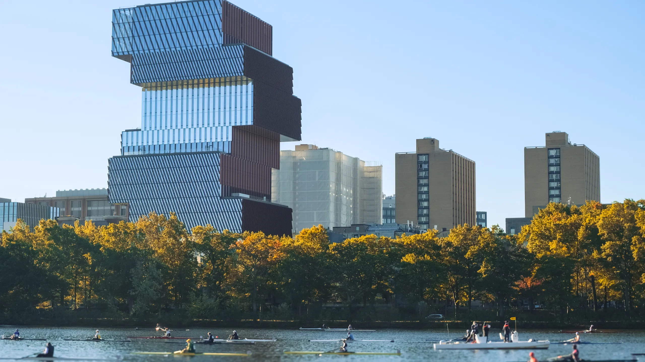 A group of unrecognizable rowers on the Charles River with Boston’s modern skyline and autumn trees in the background.