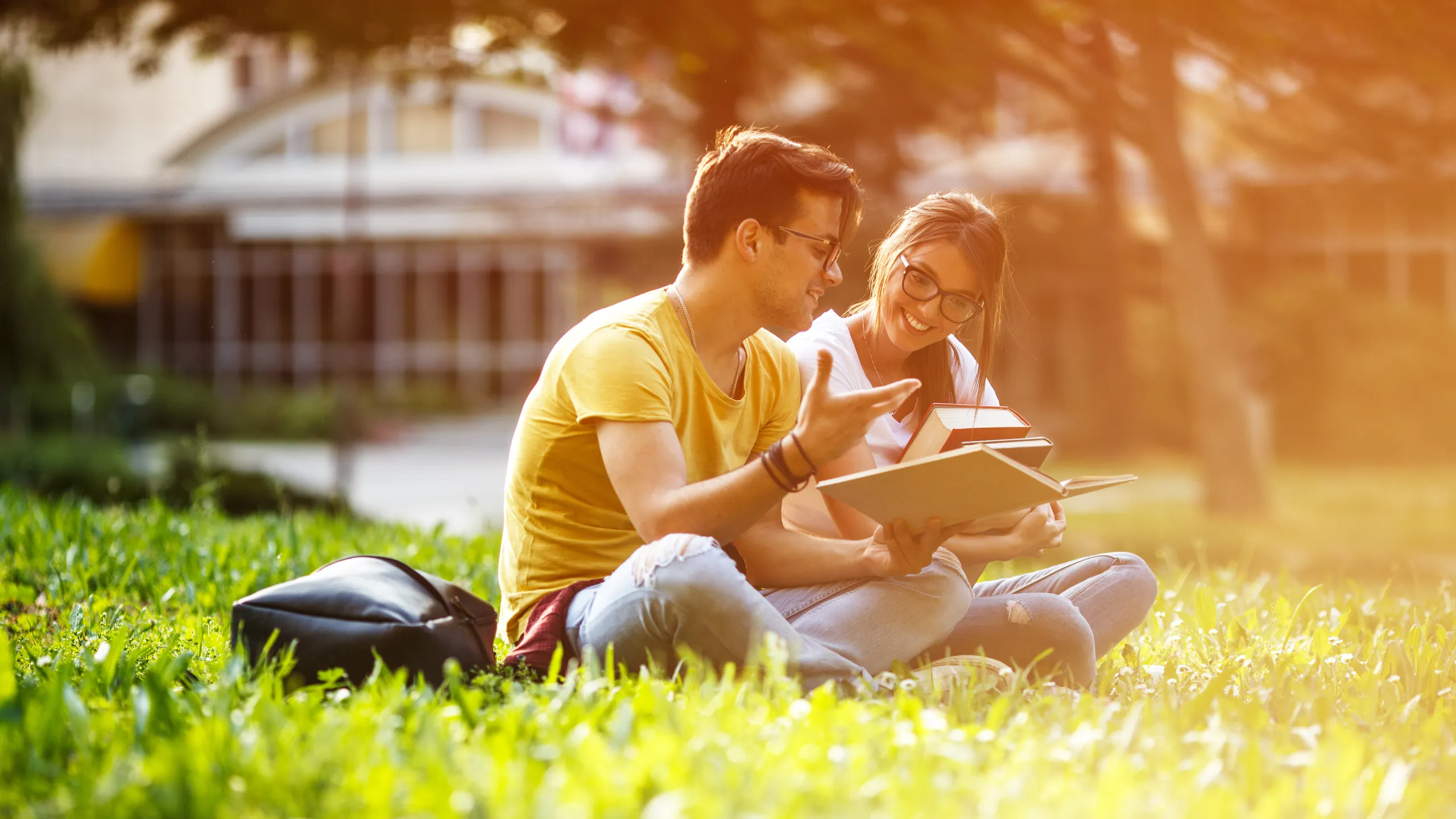 Two young students sitting at the campus yard , reading book and preparing for university exam.