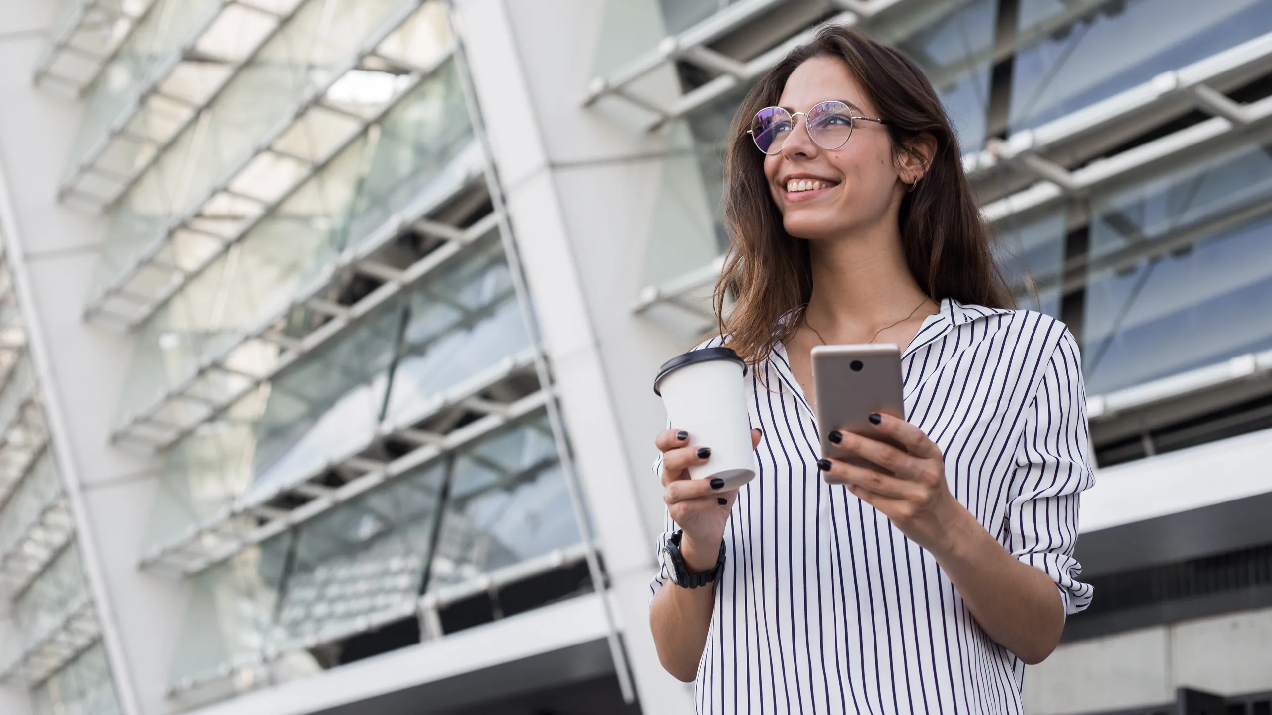 Portrait of cheerful businesswoman in eyeglasses enjoying coffee break outdoors on sunny day, prosperous trader satisfies with accomplished online payment successfully on mobile looking away