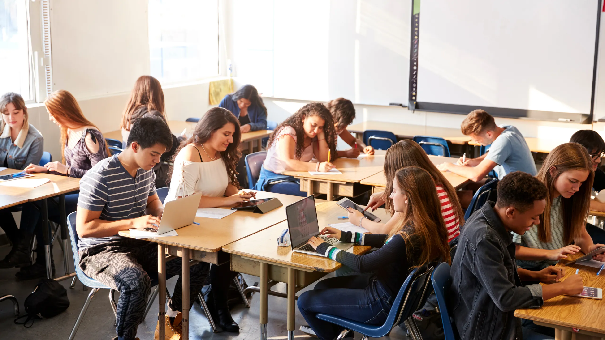 Wide Angle View Of High School Students Sitting At Desks In Classroom Using Laptops