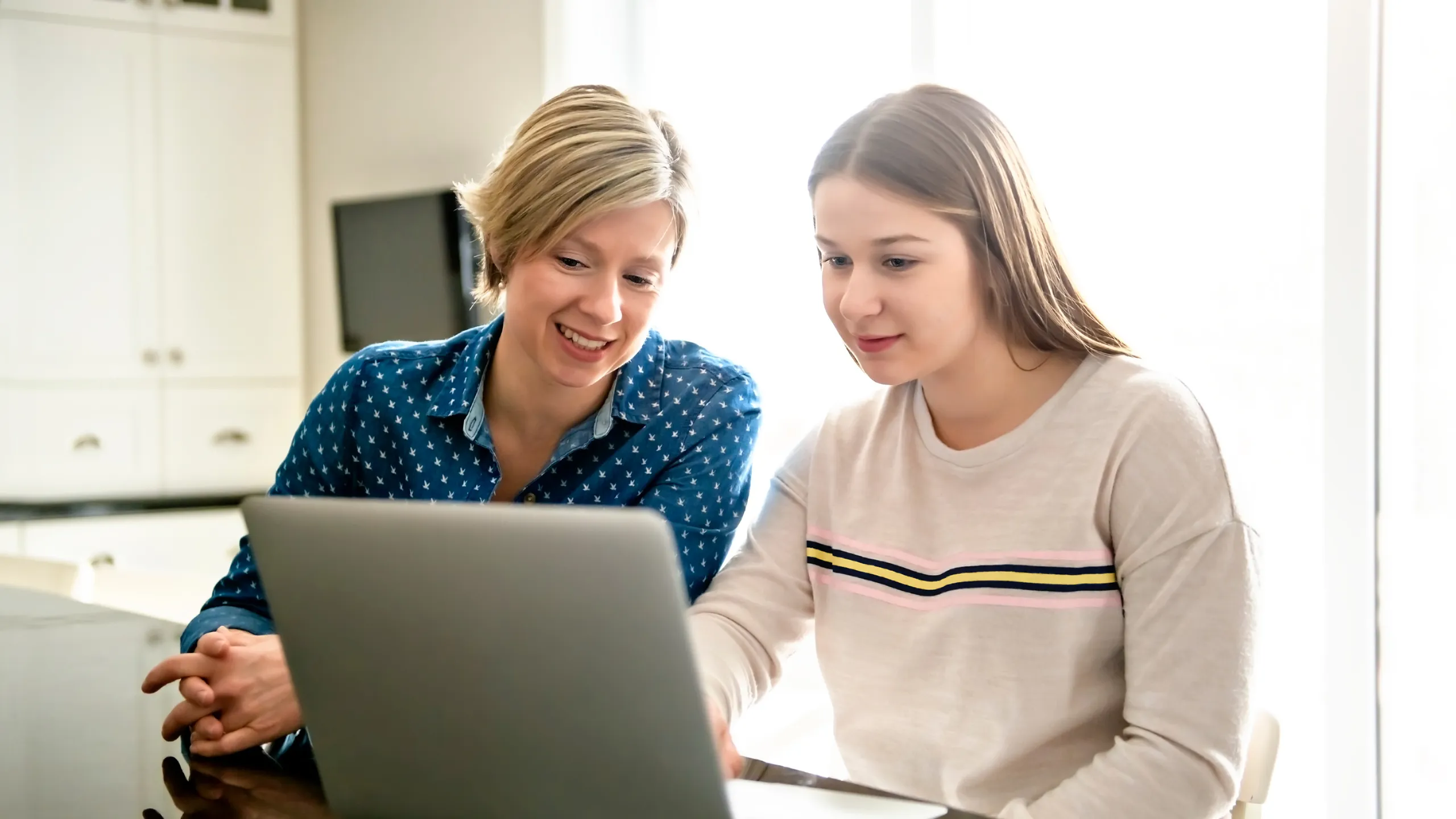 A mother using a laptop in kitchen with teenager