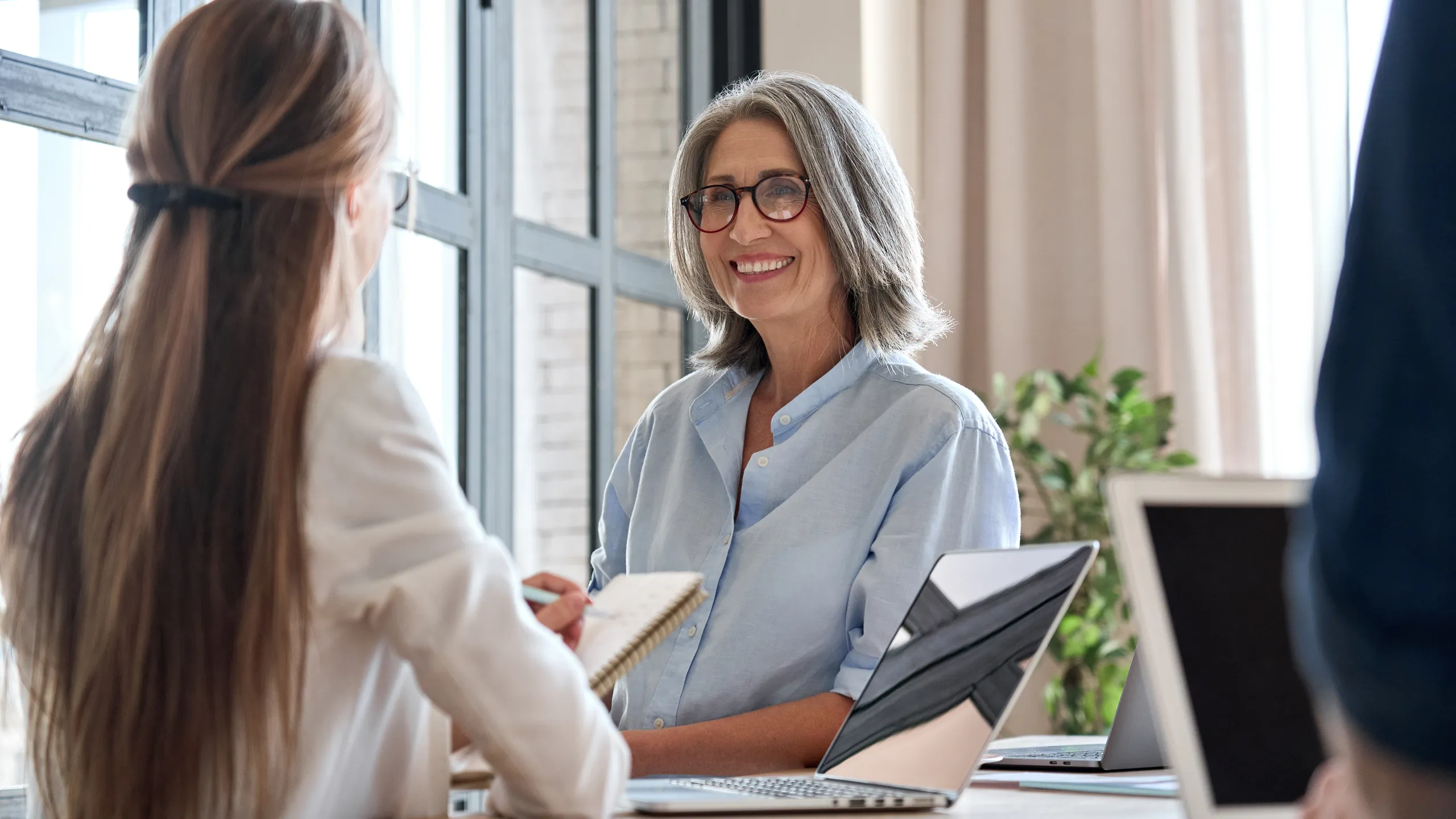 Senior business woman working on laptop with assistant writing notes.