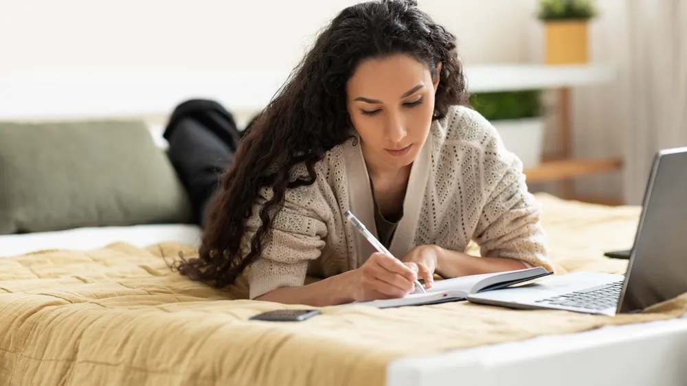 Woman lying on bed, using computer and writing in notebook