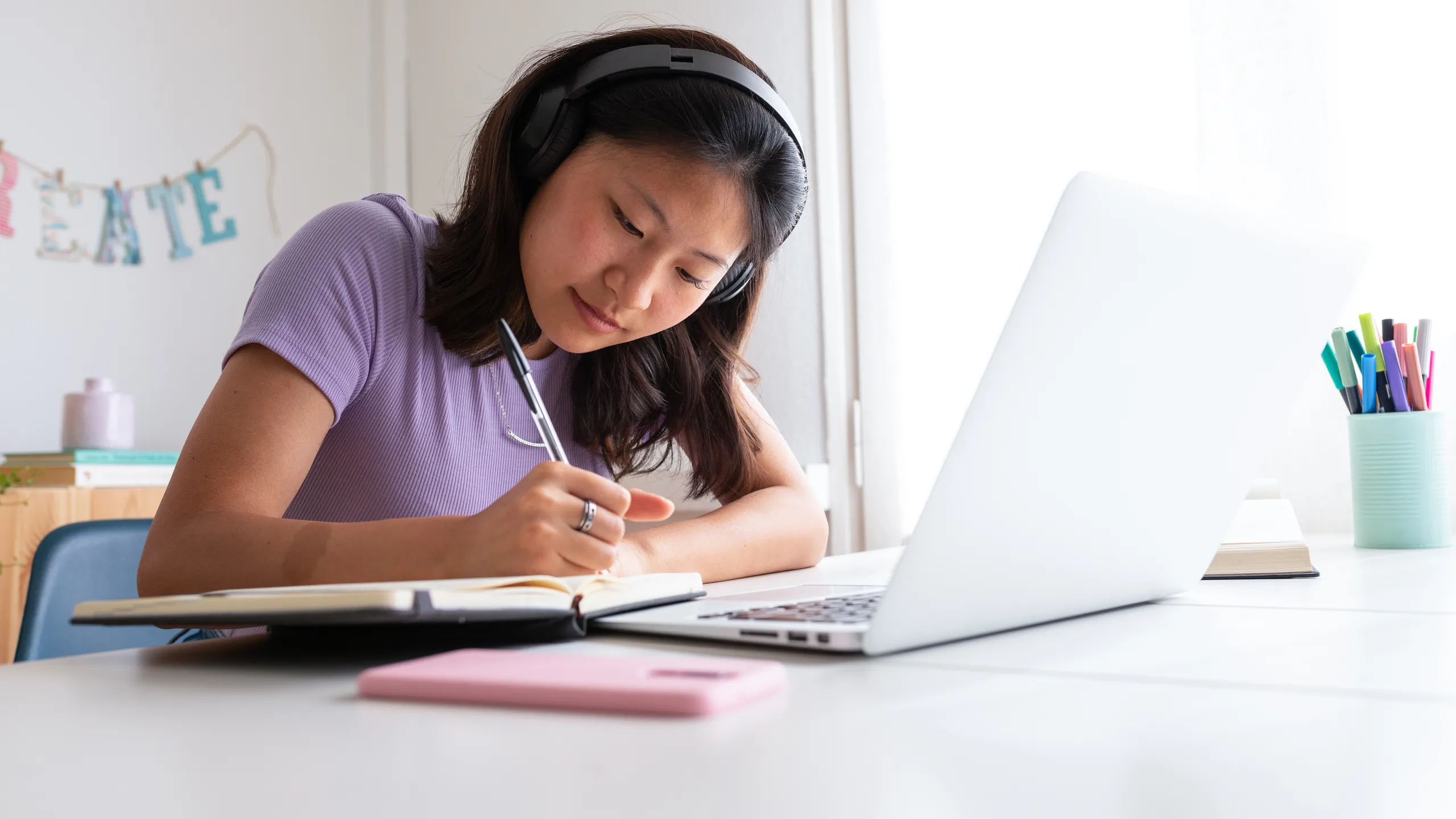 Asian female high school student writing on notebook. Chinese teen girl with headphone, studying at home using laptop.