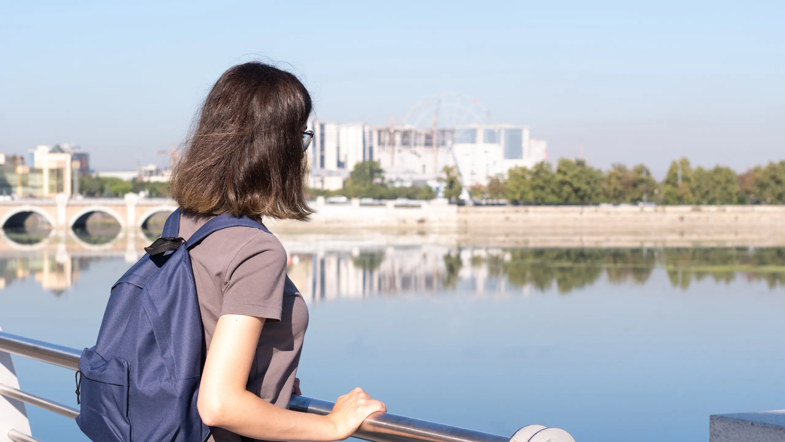 A rear view of a young curly haired college student a blue backpack Standing in a park area against the background of river