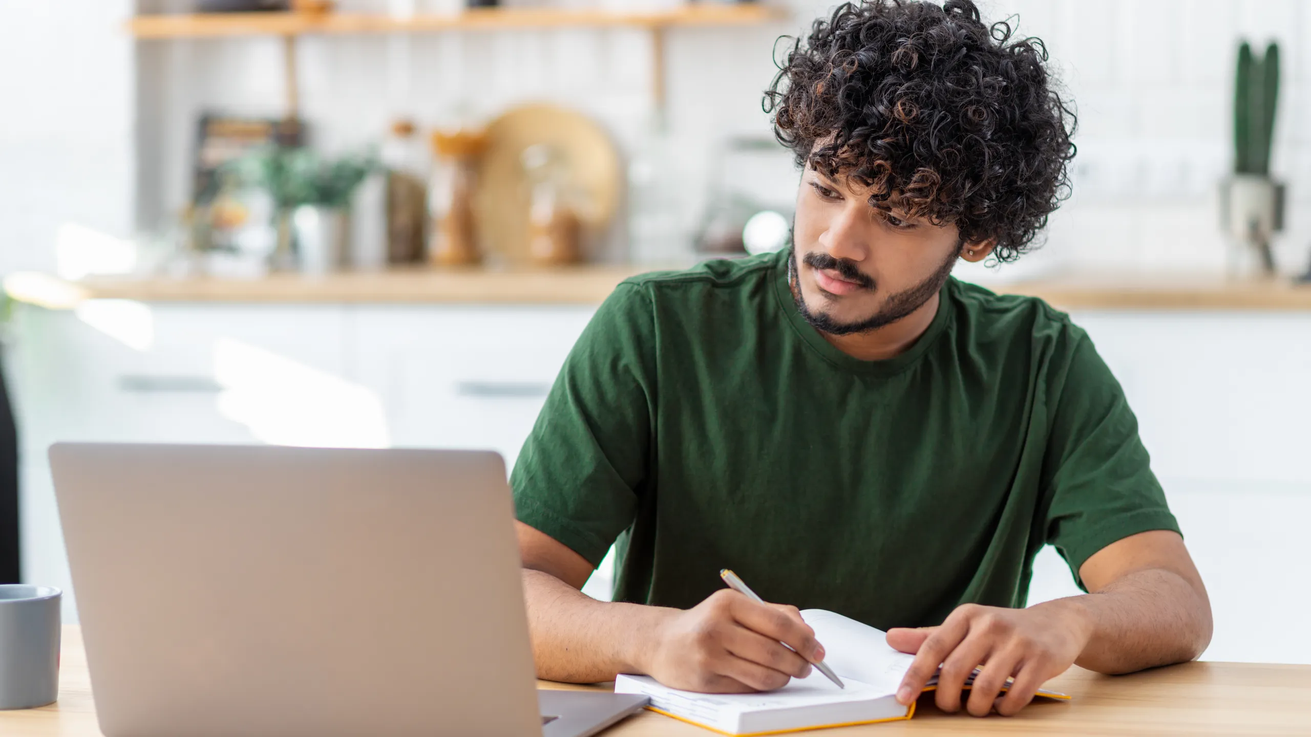 Concentrated young Asian man student using laptop computer for online education, taking notes in notebook, studying, distance learning at home