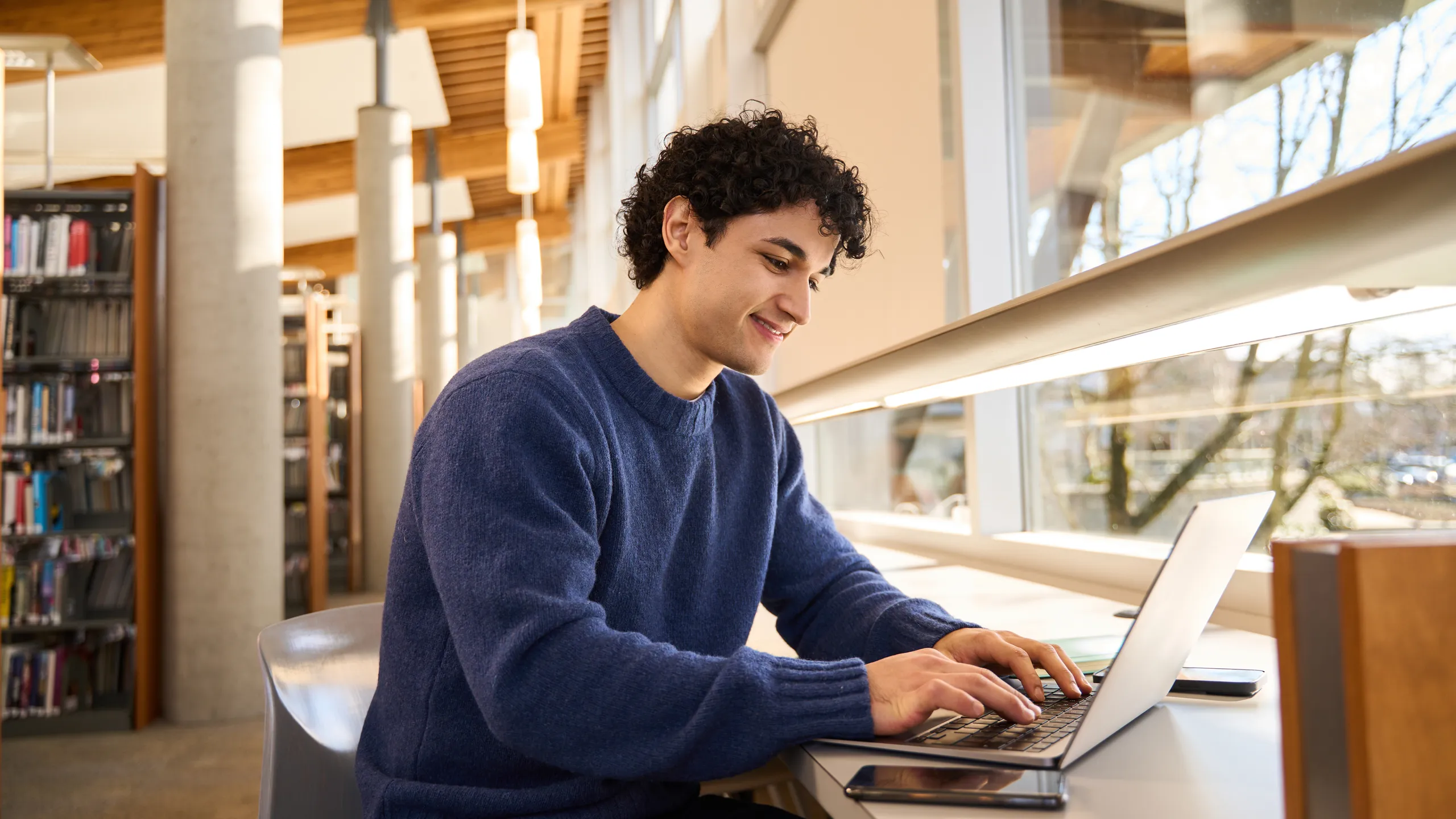 Smart Hispanic male student sitting at desk and using laptop preparing diploma project in the library campus