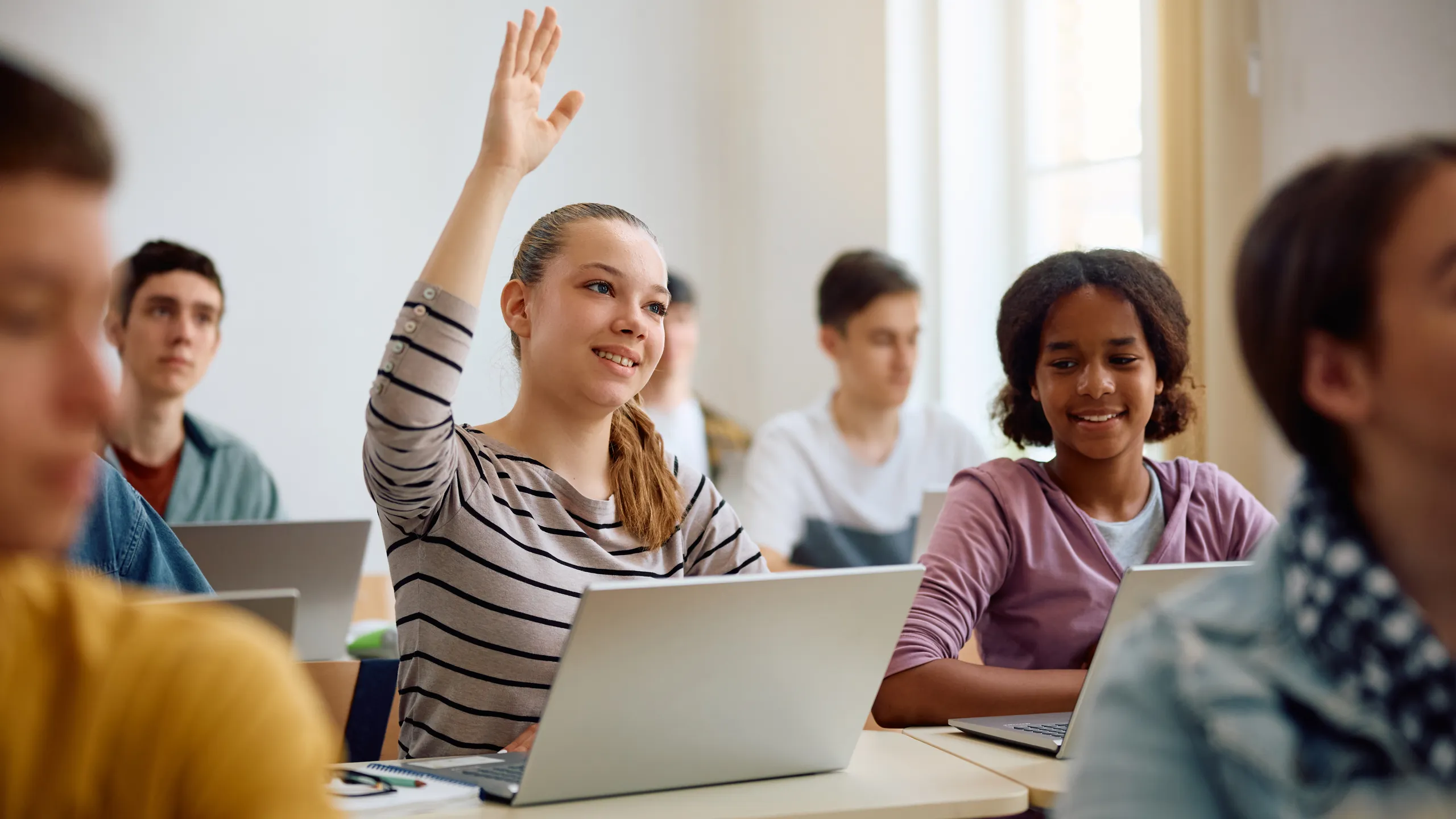 Teenage girl using laptop and raising her hand to ask question during computer class at high school.