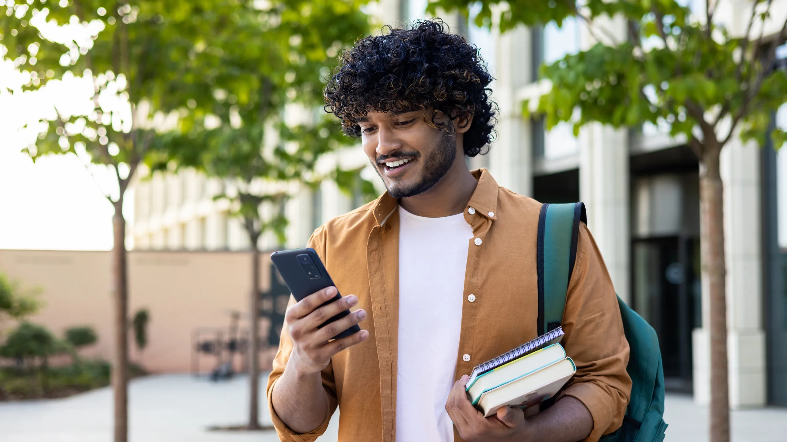 Young smiling Indian male student came to study, standing outside campus, university and using phone. He was holding books and a backpack