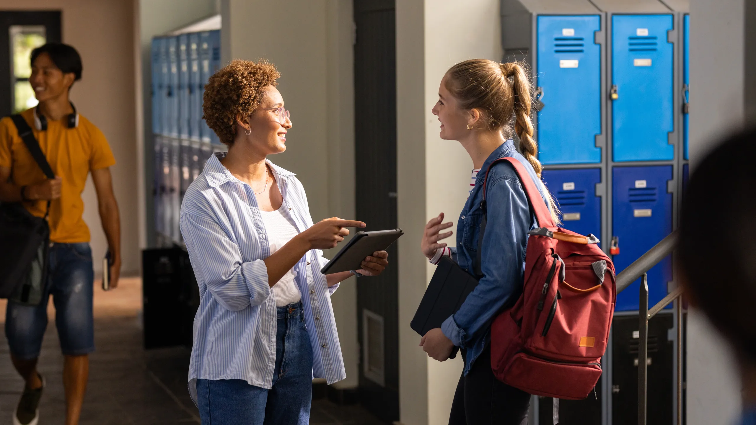 In high school, teacher with tablet talking to student with backpack by lockers
