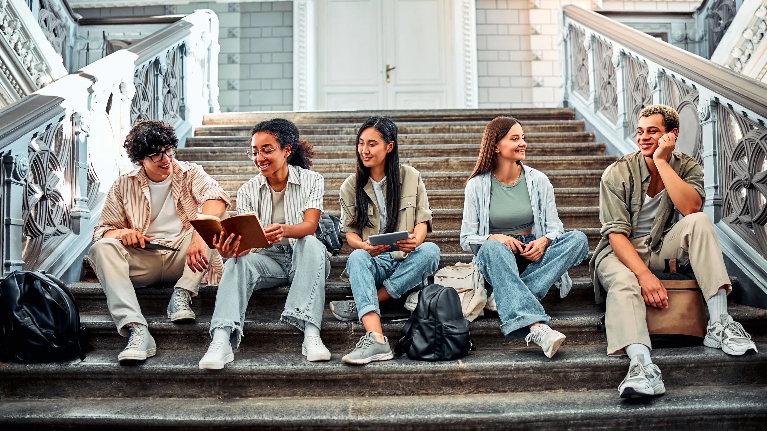 A group of modern stylish students are sitting on the steps of the university and talking, holding books, laptops and other gadgets. Student life.